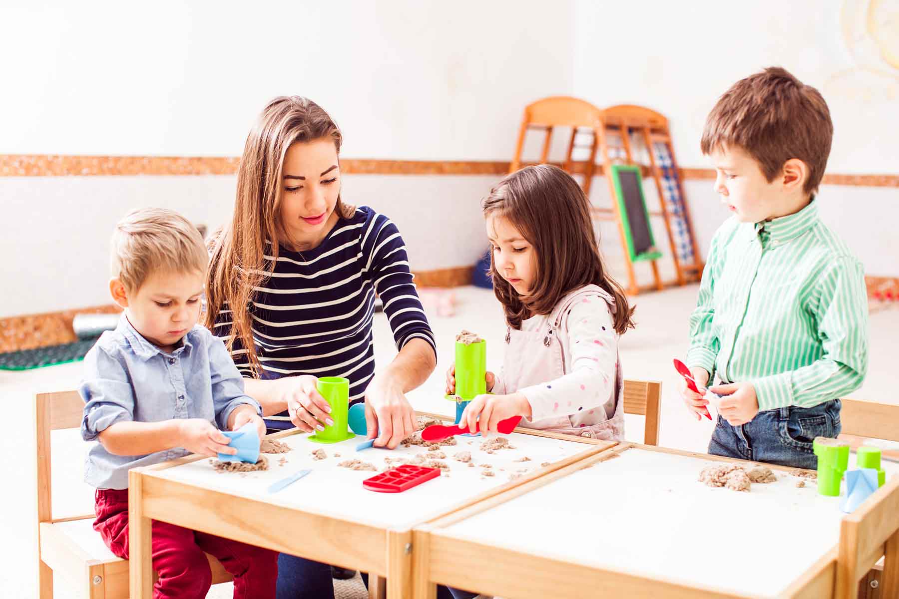 A group of young children engaged in kinetic sand activity with shaping tools alongside their teacher