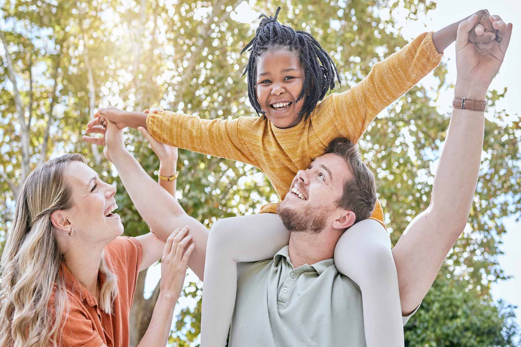 Young African American girl with foster parents, riding piggyback, smiling, and having fun outdoors, with the sun shining through the trees behind them. Young African American girl with foster parents, riding piggyback, smiling, and having fun outdoors, with the sun shining through the trees behind them.
